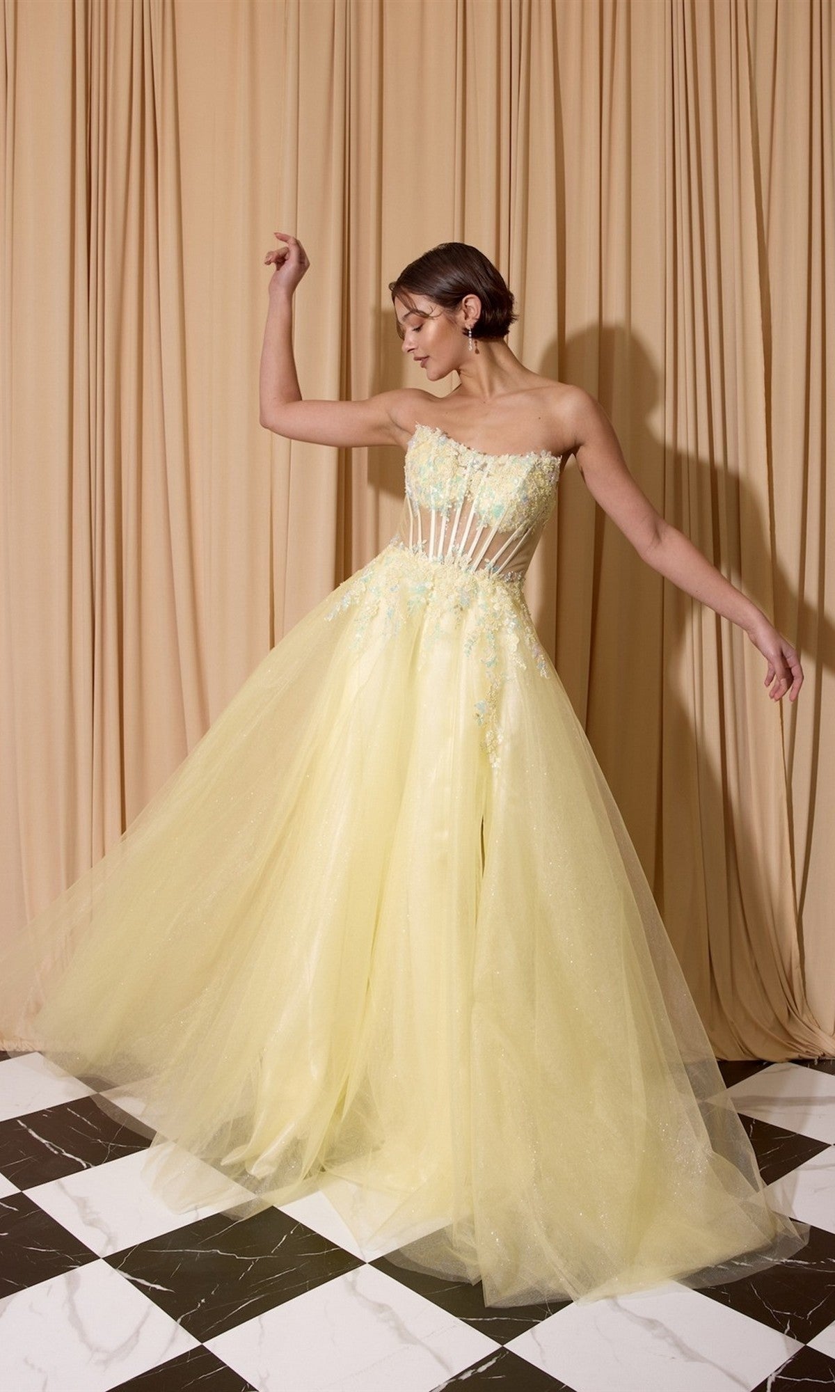 A woman in the soft yellow Long Prom Dress: Eureka EK208, featuring a strapless design with beaded floral appliqué and a sheer bodice, poses in front of beige curtains on a black and white checkered floor.